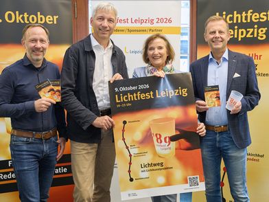 Vier Personen stehen vor Roll-ups, auf denen das Lichtfest Leipzig beworben wird. Sie halten zudem ein großes Plakat, Programmhefte und einen Kerzenbecher in der Hand.