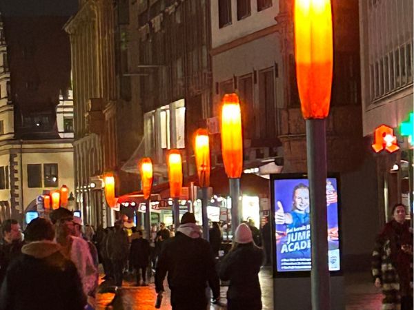 Fußgängerzone im dunkeln, Laternen sind orange beleuchtet und Menschen laufen die Straße entlang
