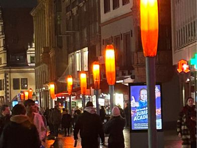 Fußgängerzone im dunkeln, Laternen sind orange beleuchtet und Menschen laufen die Straße entlang