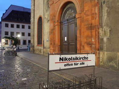 Man sieht eine große braune Eingangstür zur Kirche und davor ein Schild mit der Aufschrift "Nikolaikirche - offen für alle"